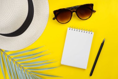 Not defteri, kalem, güneş gözlüğü, palmiye yaprağı, sarı arka planda şapka. Yaz, seyahat konsepti. Üst görünüm, düz görünüm, kopyalama alanı, maket
