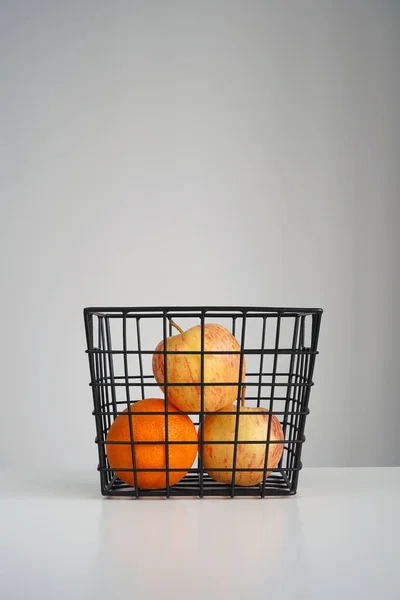 Ripe apples and orange in black grid basket on gray background. Recipe, juice, diet concept. Close-up, copy space, vertical format