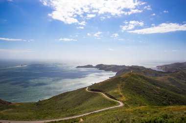 The hilly coast of the Pacific Ocean, taken from the height of the Marin Headlands. Lots of sun, green vegetation and winding road. clipart