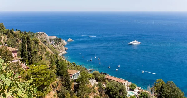 Panoramic view from Taormina, in Sicily (Italy). A sunny summer day with beach, blue sky, boats and luxury houses. A cactus can be seen among the green vegetation in the foreground. - Image