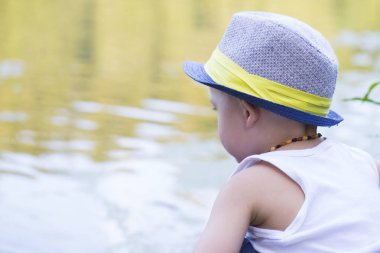 Bir şapka ile şirin küçük bir çocuk portresi nehir kenarında oturan ve güzel ve güneşli gün keyfini çıkarın. Yaz günbatımı renkleri ve atmosfer. Kopya girintili