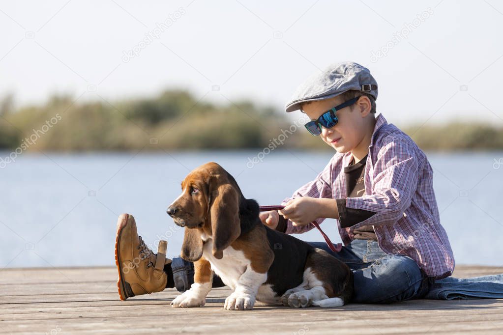 Little sweet boy with a hat and sunglasses sits by the river with his dog. They enjoy together on a beautiful sunny day. Child hugging his dog. Growing up, love for animals - dogs, free time, travel, vacation. Copy space 