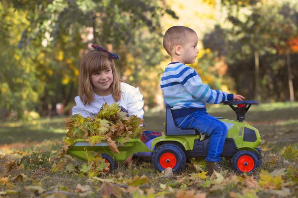 小男孩和一个女孩在外面玩耍 他们在大自然中享受一个美丽的阳光明媚的秋天 小兄弟和妹妹喜欢一起度过时光 一个小男孩骑着他的小拖拉机 女孩收集落叶 — 图库照片