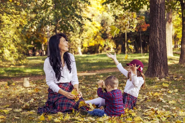 美丽的年轻母亲在公园里和孩子们玩耍 他们在玩干落叶的时候很开心 — 图库照片