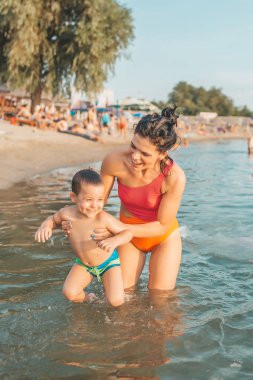 Üç yaşındaki yürümeye başlayan çocuk annesi su ile oynamak. Mutlu anne küçük çocuk kucağında tutarak. Mutlu bir aile. Tatil, seyahat kavramı