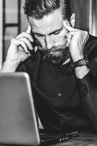 Young bearded sad businessman is sitting at table, covering his face with his hand and talking on cell phone. On desk is laptop, tablet computer. Man got bad news.Stress. Black and white photo