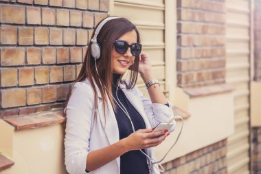 Güneşli bir günde sokakta kulaklık ile müzik dinlemek mutlu güzel ve genç birbayan. Tuğla duvar arka plan