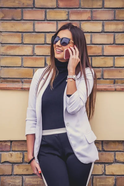 Üstünde hareket eden telefon yanında kahverengi tuğla duvar açık havada güneşli günde konuşurken gülüyor güneş gözlüklü genç mutlu womanl