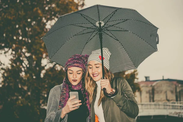 Δύο Νεαρές Γυναίκες Ομπρέλα Και Χρήση Κινητού Τηλεφώνου Στην Πόλη — Φωτογραφία Αρχείου