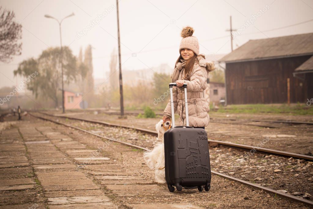 Happy little girl with her dog and suitcase on a railway station. Kid with dog waiting for train and happy about traveling.