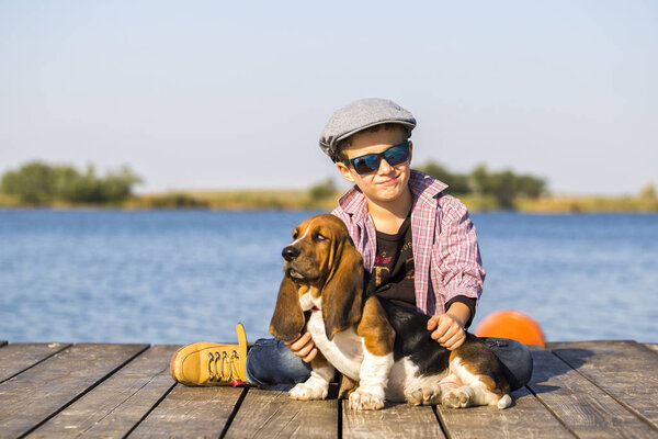 Little cute boy is sitting by the river with his dog. They enjoy together on a beautiful sunny day. Boy hugging his puppy. Growing up, love for animals - dogs, free time, travel, vacation. Copy space