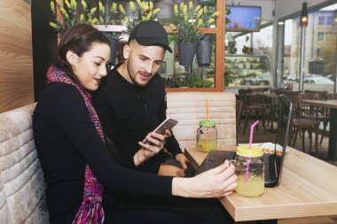 Mutlu genç çift kafede otururken bir dizüstü bilgisayar ve cep telefonu kullanıyor. Smartphone kullanarak ve gülümseyen kafeteryada oturan iki mutlu arkadaş .