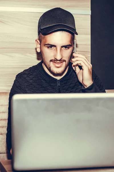 Young man with cap talking on the phone, using laptop computer and drinking coffee in a coffee shop