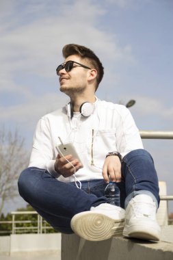 Yakışıklı genç adam oturur ve açık havada sahiptir. İnternette surf ve müzik dinlemek. Seçici odak ve küçük alan derinliği.