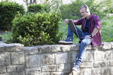 Yakışıklı genç adam parkta oturan tablet kullanmak ve müzik dinliyor. Seçici odak ve küçük alan derinliği.