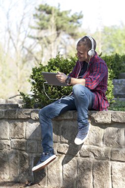 Yakışıklı genç adam parkta oturan tablet kullanmak ve müzik dinliyor. Seçici odak ve küçük alan derinliği.