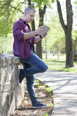 Yakışıklı genç adam stading parkta, tablet kullanmak ve açık havada. Seçici odak ve küçük alan derinliği.