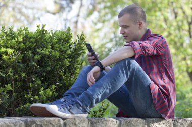 Parkta oturan ve telefon tutarak yakışıklı bir genç adam. Seçici odak ve küçük alan derinliği.