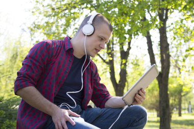 Yakışıklı genç adam parkta oturan tablet kullanmak ve müzik dinliyor. Seçici odak ve küçük alan derinliği.
