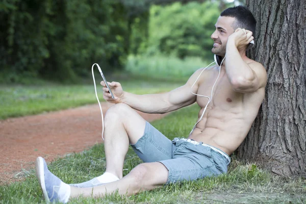 Handsome muscular young man sits in a park next to the tree, relax and listen to music