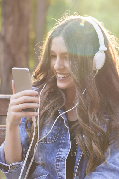 Beautiful young smiling woman sitting on a park bench, listening to music and holding a mobile phone. Lens flare.