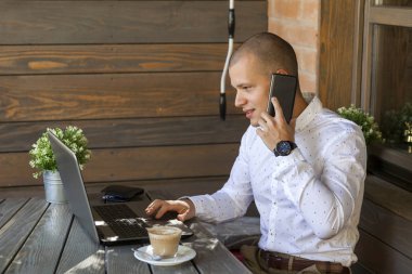 Yakışıklı genç adam oturur, laptop görünüyor, sörf internet ve kahve içme. Genç işadamı, iş ortaklarıyla cep telefonda konuşurken