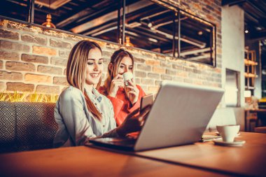 Dizüstü bilgisayar kullanarak ve bir modern café iç otururken kahve içme iki gülümseyen kız. Dostluk ve teknoloji. Seçici odak ve küçük alan derinliği.