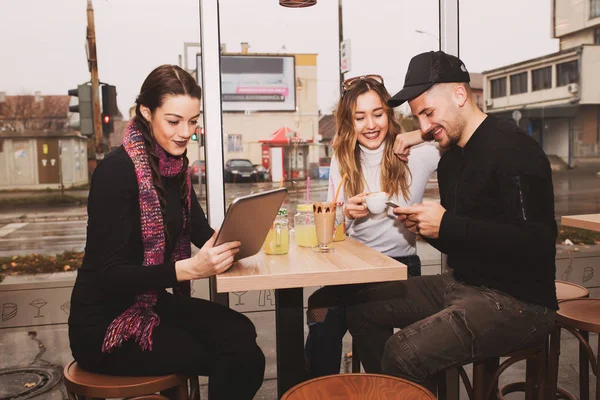 Cheerful group of friends, one man and two women sitting at the table in the cafe shop,  talking and having fun while using smart phone and tablet.