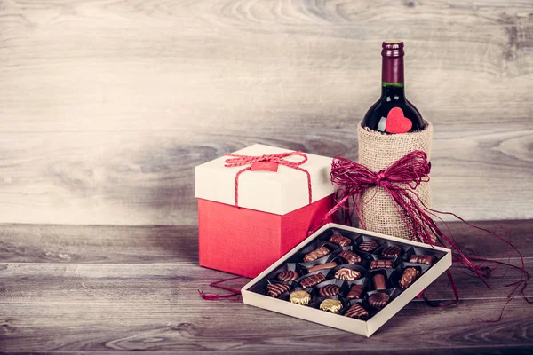 Red wine bottle, box of chocolates and gift with decoration by red heart on wooden table. Valentines day celebration concept. Copy space.