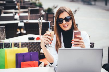 Mutlu genç iş kadını Sokak kafede masada oturan ve alışveriş sonra kahve yudumlarken laptop ve smartphone kullanarak güneş gözlüklü. Kız alışveriş online, bloglama, sohbet.