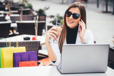 Sokak kafede masada oturan ve cep telefonuyla konuşurken ve alışveriş sonra kahve içme sırasında dizüstü kullanarak mutlu genç iş kadını. Kız alışveriş online, bloglama, sohbet.