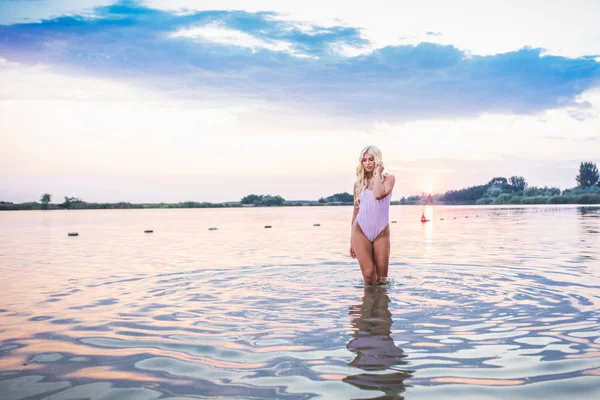 Portrait of young stunning woman in swimsuit standing in lake. Beautiful girl posing in water at sunset