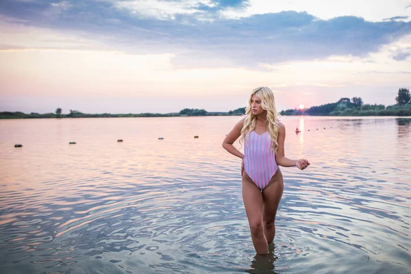 Portrait of young stunning woman in swimsuit standing in lake. Beautiful attractive girl posing in water at sunset