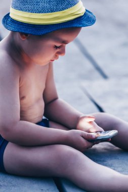 Sevimli küçük yürümeye başlayan çocuk kumsalda oturuyor ve smartphone ile oynamak şapka ile. Çocuk akıllı telefon kullanmayı öğrenmek. Çocuk manifatura telefon. -teknoloji ve yaşam tarzı kavramı