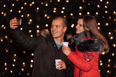 Selfie yapma ve mulled şarap fincan tutarak ve önünde bir Noel arka plan - IMAG ayakta iken gülümseyen mutlu genç çift