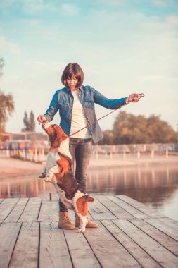 Elini tutuyor ve onu köpek Basset Hound ayakta nehrin yanında arka ayakları üzerinde oynamaktan genç kadın görünümünü