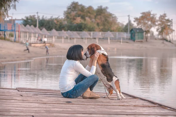 Portrait of beautiful young woman sitting next to the river and hugging her dog Basset Hound. Happy woman likes to spend time with her dog.