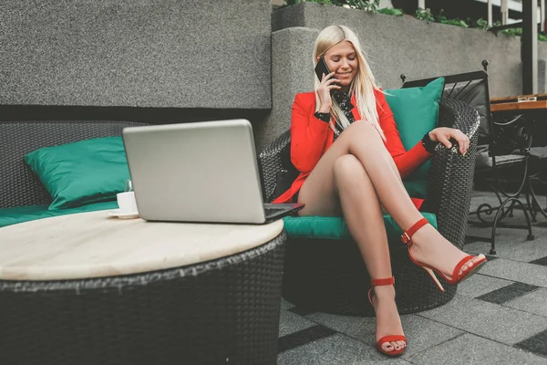 Gorgeous blonde young business woman having cell phone conversation while sitting front open laptop computer in outdoor coffee shop, female person talk on smart phone - Image