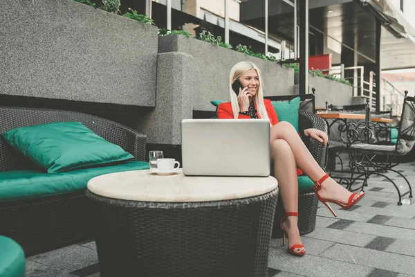 Attractive blonde young business woman having cell phone conversation while sitting front open laptop computer in outdoor coffee shop, female person talk on smart phone - Image