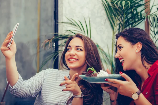 Two young women eating cakes and taking selfie with smartphone