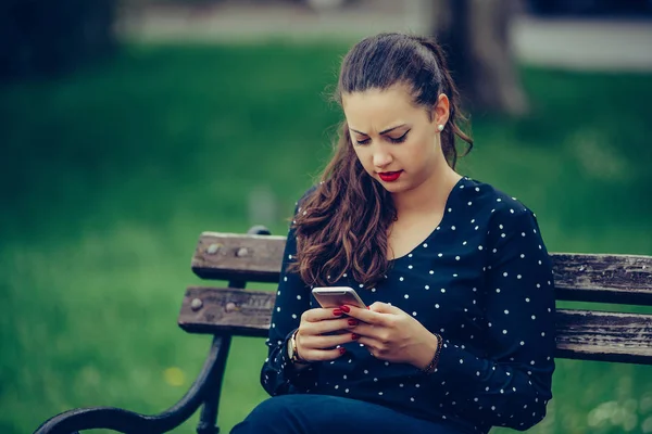 Девушка пишет смс на смартфоне, сидя на скамейке в парке — стоковое фото