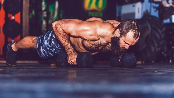 Muscular bearded man doing push up exercise with dumbbell in gym