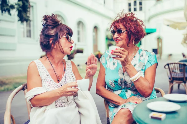 Two happy senior women talking and drinking coffee while looking — Stock Photo, Image