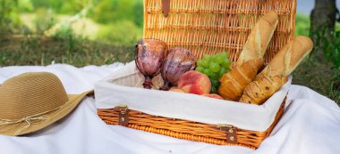 Çimlerin üzerinde piknik sepeti, üzerinde yiyecek ve battaniye var. Güneşli bir günde bir tarlada piknik öğle yemeği. Baget, kruvasan, şeftali, bir buket kır çiçeği ve üzüm. Fransız tarzı piknik