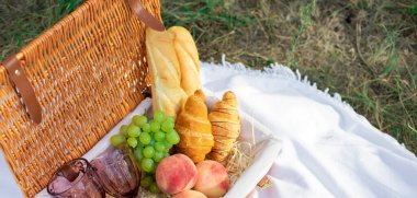 Çimlerin üzerinde piknik sepeti, üzerinde yiyecek ve battaniye var. Güneşli bir günde bir tarlada piknik öğle yemeği. Baget, kruvasan, şeftali, bir buket kır çiçeği ve üzüm. Fransız tarzı piknik