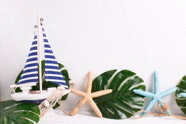 Yaz, tatil kavramı. Dekoratif ahşap oyuncak tekne, deniz yıldızı ve tropikal bitki yaprakları beyaz dokulu arka plan üzerinde. Metin için yer. 