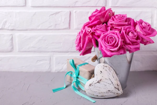 Bright pink roses flowers in bucket, heart  and wrapped box wih present on grey background against white wall. Selective focus. Place for text. Floral still lif