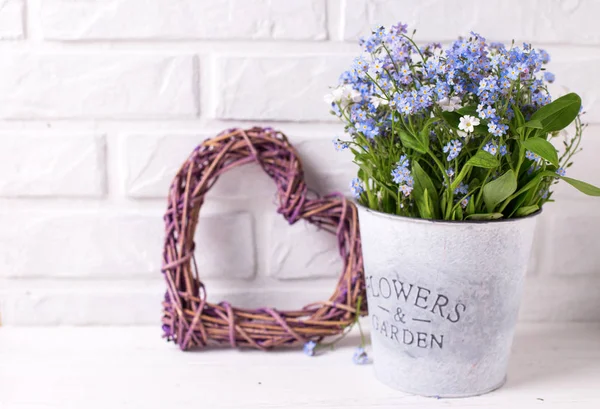 blue forget-me-nots or myosotis flowers in grey bucket, decorative heart and pearls on white wooden background