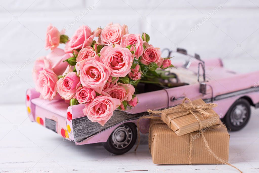 Retro car toy with pink roses and wrapped boxes with presents flowers against white textured wall