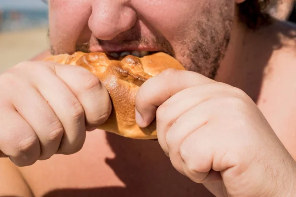 Fat man on the beach eating a pie. Overweight and unhealthy diet